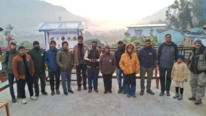 A group of approximately 15 people, a mix of diverse ages and backgrounds, posing for a group photograph at The Corbett Rajae Homestay. They are standing in front of a banner that reads "KALAGARH NATURE FESTIVAL," featuring wildlife and nature logos. The setting is a terrace overlooking a scenic valley with terraced farms and distant mountains. It is dawn or early morning, indicated by soft lighting and some mist.