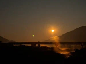 A wide-angle, vibrant, late-afternoon photograph taken from an elevated overlook. Two men are seated on a blanket on the ground in the foreground (backs to the camera), looking out over a massive, hazy valley. Sitting beside them is a golden-brown dog, also looking out. The valley below contains a river, terraced farms, a small village, and a distant bridge. The background is composed of multiple layers of green and blue-grey mountains under a dramatic orange-to-blue sky. Foreground elements (like plant leaves) are sharply focused.
