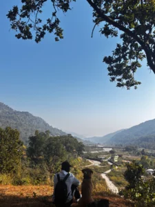 A back view of a traveler with a backpack and his dog, seated on a ledge overlooking a wide, sun-drenched valley with a village and river in North Jim Corbett, under a clear blue sky.