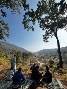 A group of approximately six adults (backs or sides mostly to the camera) are seated on a striped blanket on a grassy hillside. They are at an elevated viewpoint looking down into a vast valley during midday. The valley features a large cluster of low-lying village buildings and farms, leading to distant mountain ranges. The foreground is framed by large, overhanging oak trees with many leaves. The sky is bright, clear blue with a slight haze. One person stands at the edge of the hill.