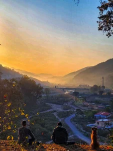 A silhouette photograph of a bridge spanning a river, set against a dramatic, hazy, orange-to-dark-blue sunset sky. The scene is heavy with mist or fog rising from the river and valley, and the surrounding mountains are visible as large, dark shapes on either side. A single, distinct, orange lens flare (the sun) is prominent, and another small, green flare is visible.
