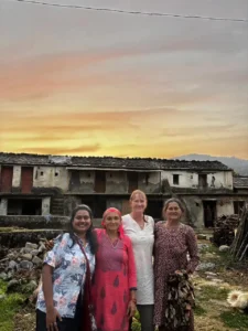 A foreign traveler and a young woman smiling with local village women in front of a traditional stone house in Uttarakhand.